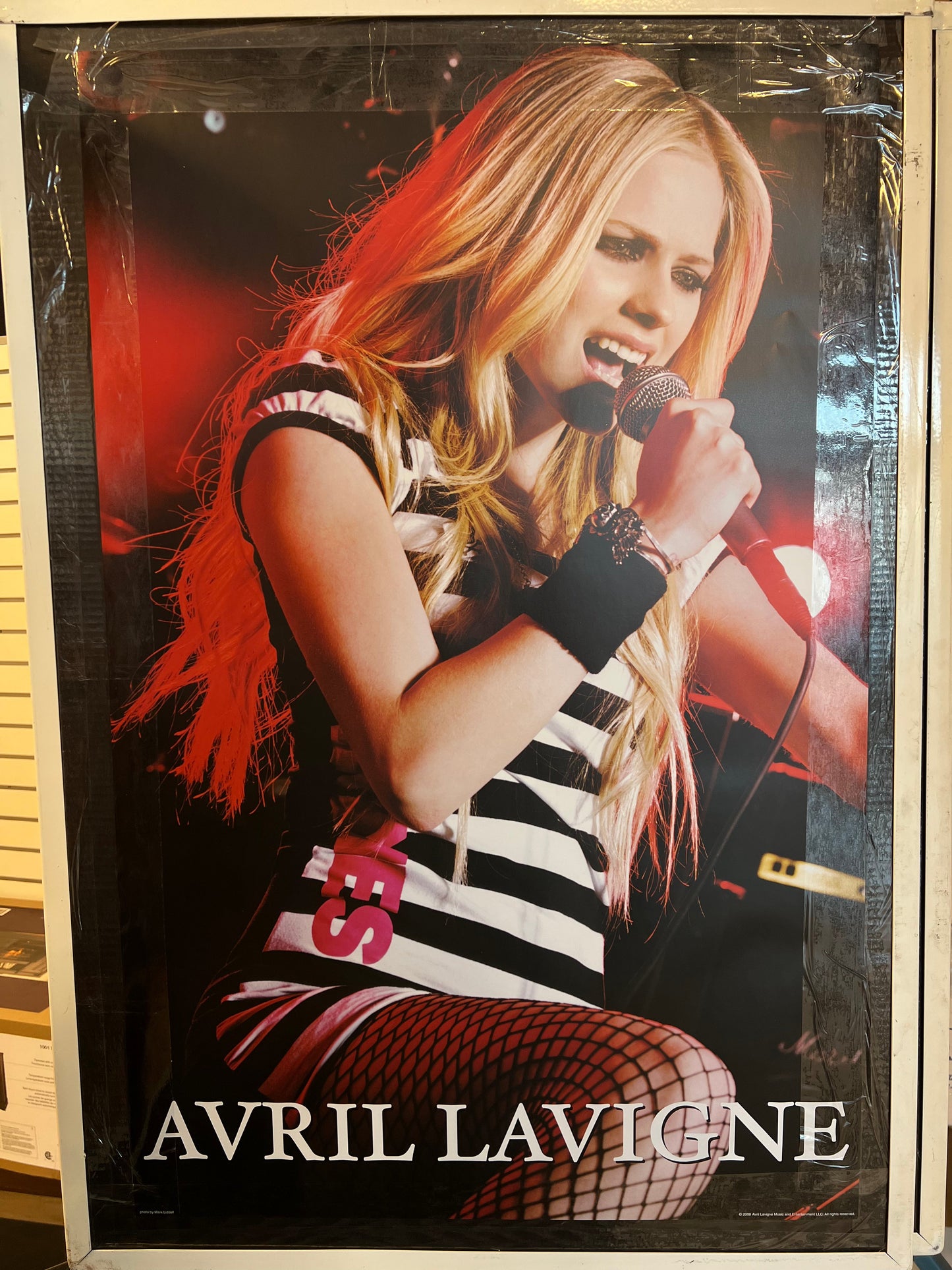 limited edition 24x36 inch vintage Avril Lavigne poster featuring the iconic image of Avril performing on stage, wearing a black and white striped mini dress, fishnet stockings, and holding a Shure microphone. The poster is printed in mint condition and will never be printed again, making it a rare and valuable piece of music history.